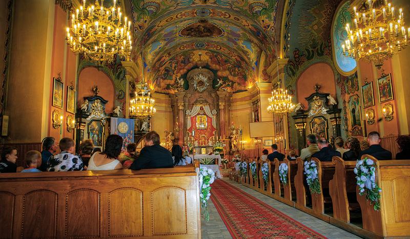Wnętrze drewnianego kościoła. Bogato polichromowany strop, kryształowe żyrandole, złoty ołtarz główny z cudownym obrazem, ołtarze boczne i drewniane ławki po bokach z siedzącymi ludźmi.