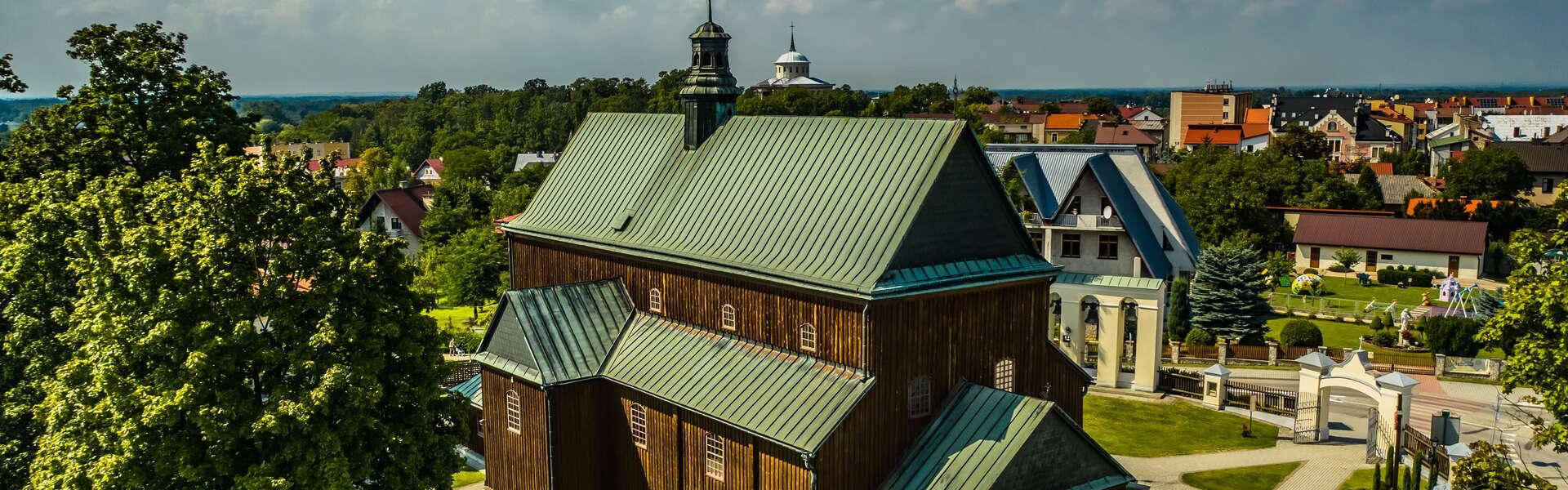 Blick auf die Holzkirche in Dąbrowa Tarnowska. Das Gebäude ist von Bäumen umgeben. Im Hintergrund Stadtbebauung.