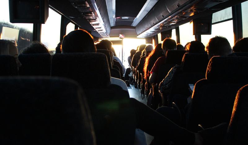 Zdjęcie ukazujące wnętrze autokaru wewnątrz którego siedzą ludzie. Ciepłe refleksy słoneczne wlewające się przez przednią szybę do wnętrza pojazdu tworzą ciekawą kompozycję zaostrzając sylwetki podróżujących.
