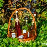 Zdjęcie przedstawia na pierwszym planie wiklinowy koszyk w którym znajdują się butelki z różowym winem pochodzącym z winnicy zawisza. W tle rosnące krzewy winorośli na których dostrzec można owoce koloru granatowego.