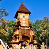 Drewniana, wysoka, smukła, bogato zdobiona kaplica na cmentarzu. Za nią drzewa w kolorach jesieni, przed nią drewniane krzyże.
