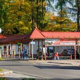 Budynek dworca autobusowego, przed którym ustawione są drewniane ławki, na których siedzą ludzie.