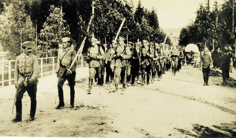 Historyczne zdjęcie w sepii - kolumna idących żołnierzy ze strzelbami. Po obu stronach drogi drzewa.